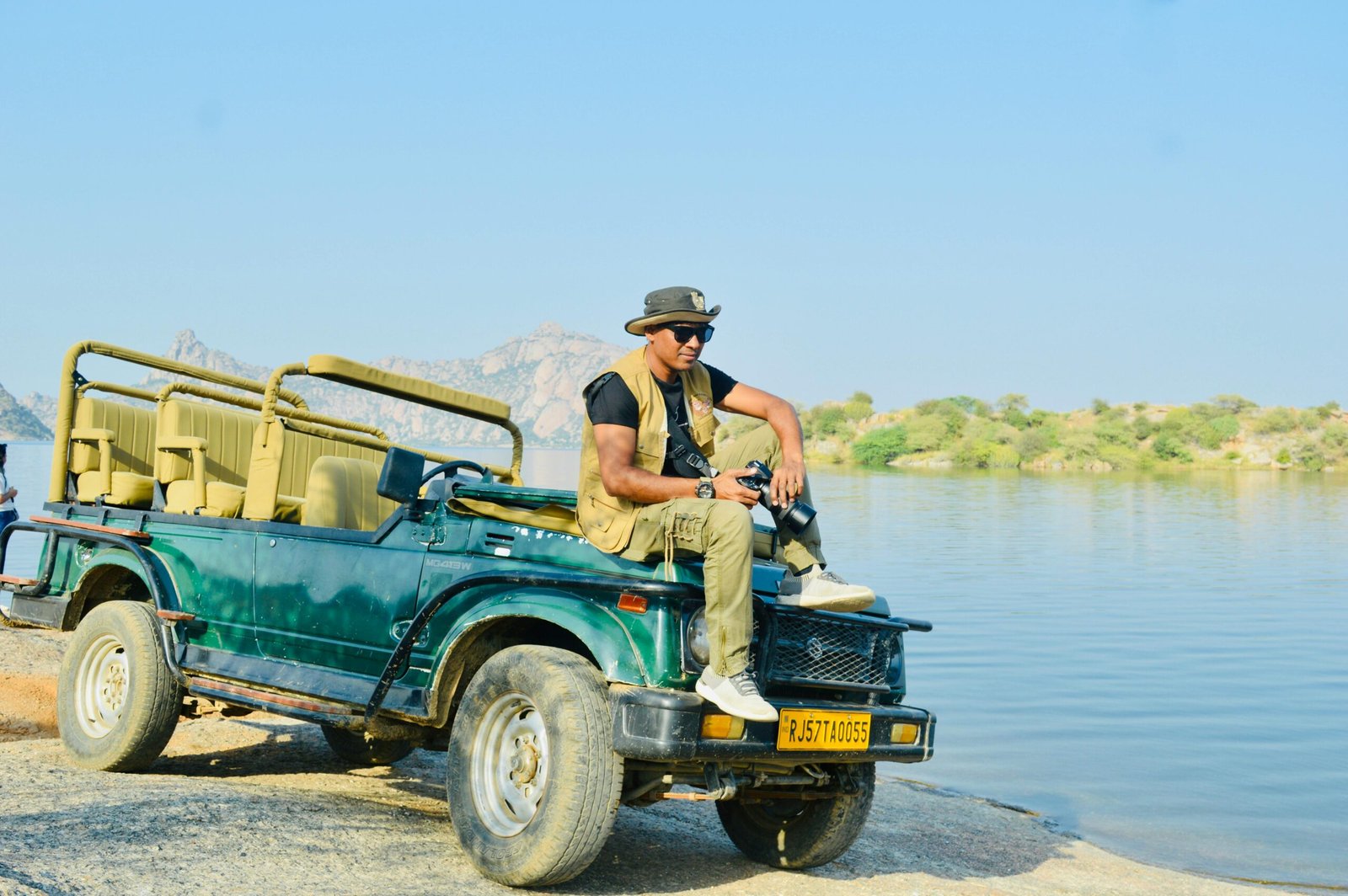 A man in safari gear sits on a jeep near a serene lake in an arid landscape.